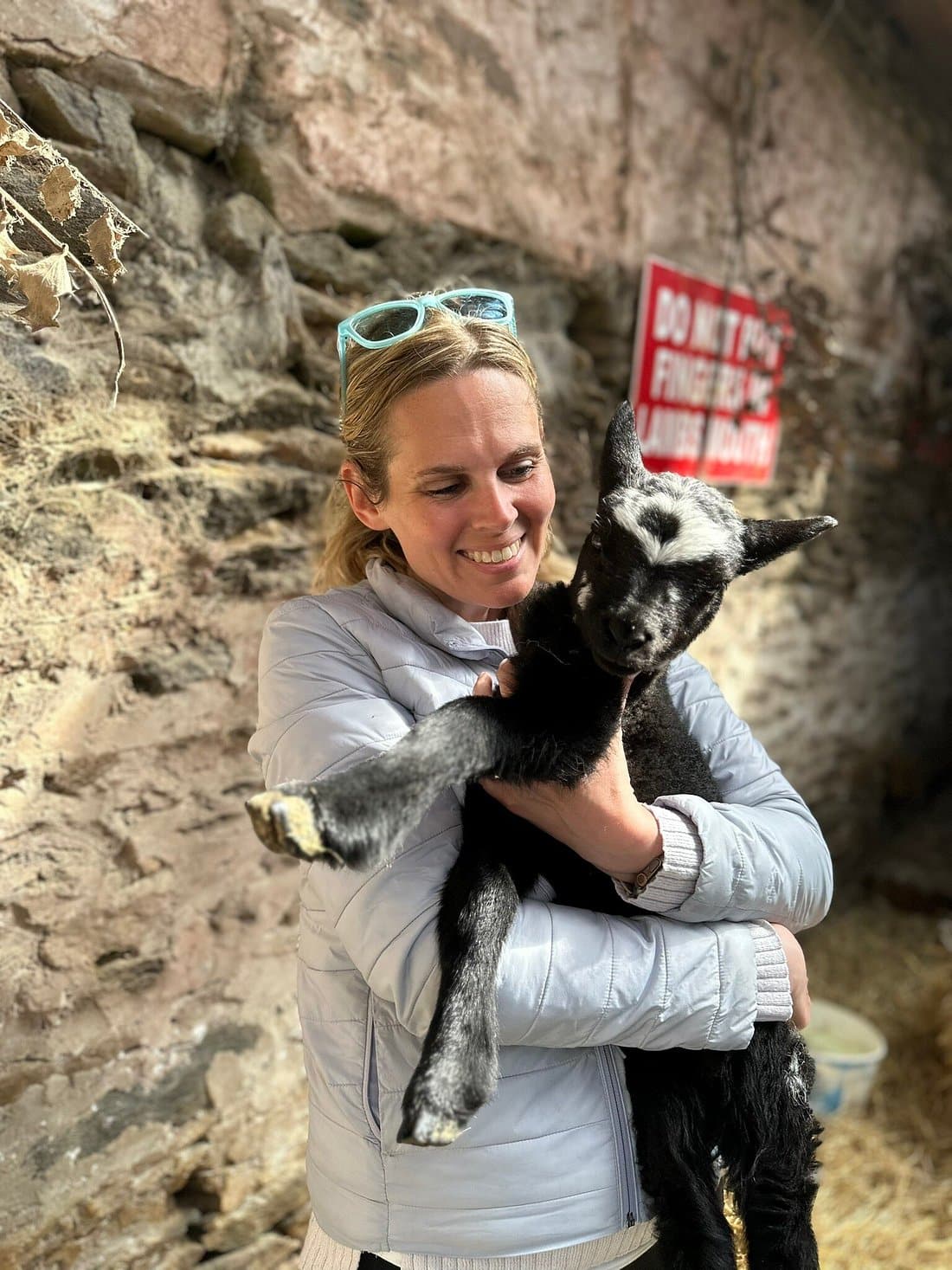 Woman smiling and holding a young goat in a rustic barn setting.