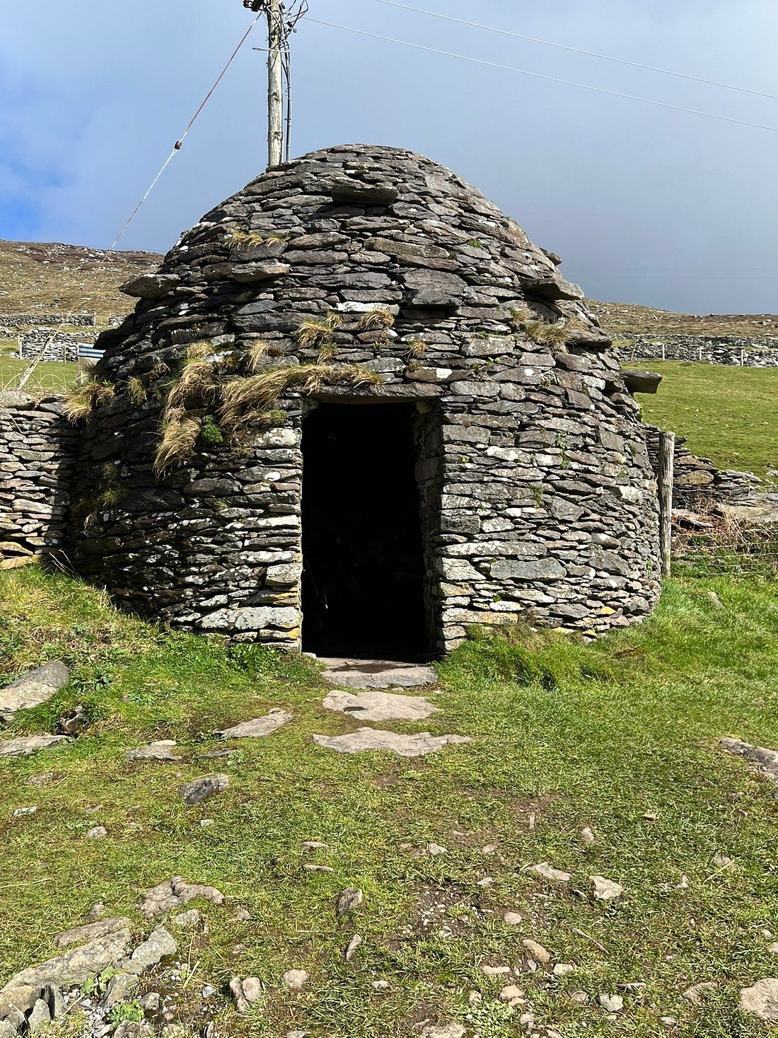 Round stone hut with a grass roof in a grassy landscape under a cloudy sky.